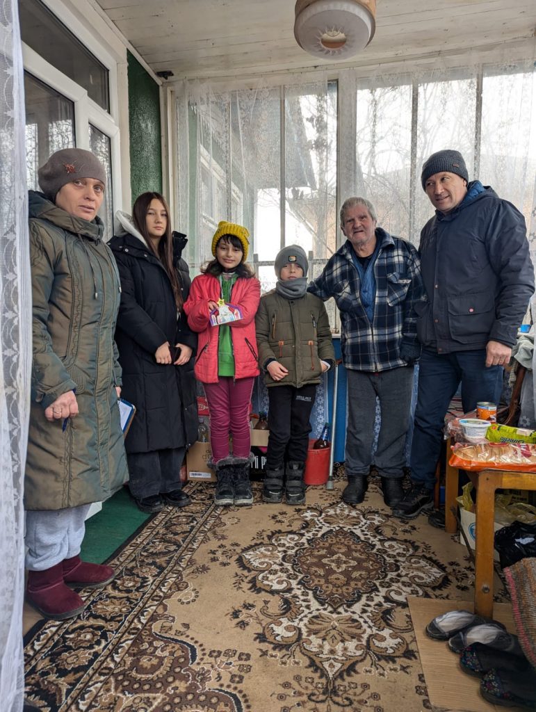 Victor, a volunteer, and two children posing with an elderly beneficiary in his home.