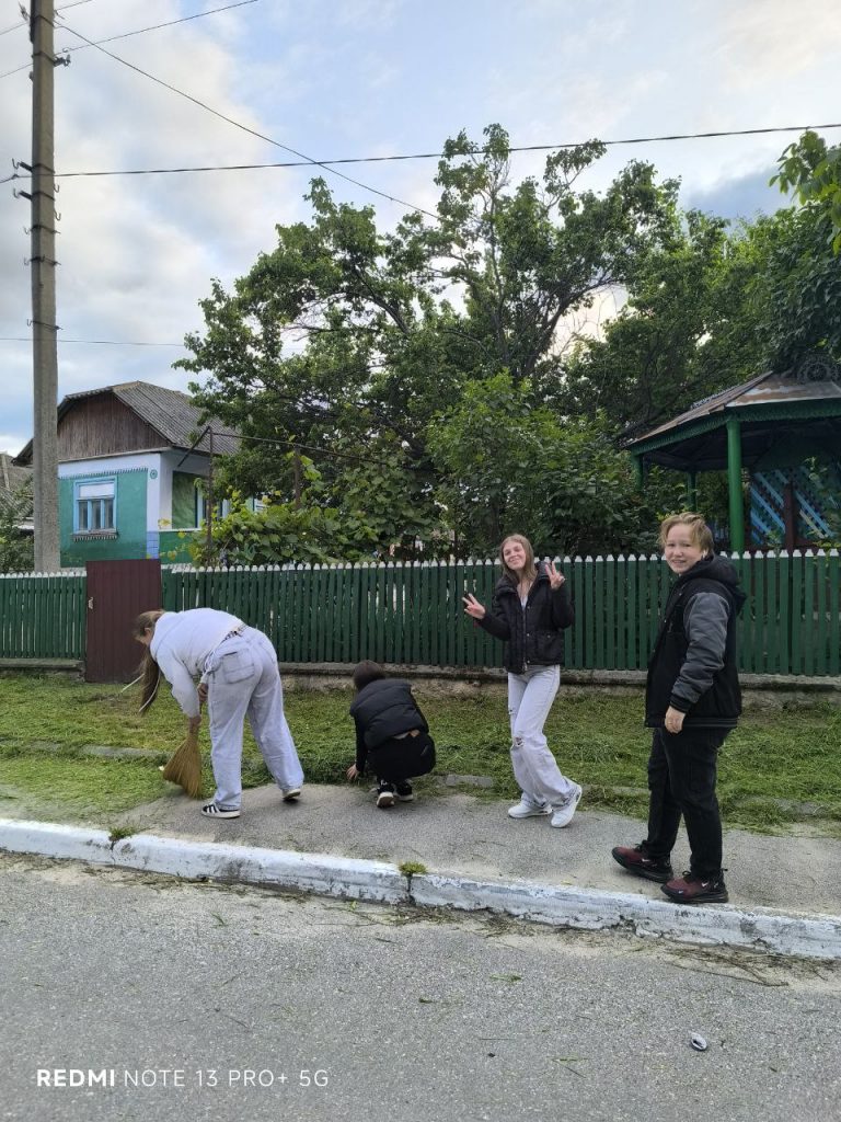 Four volunteers on a sidewalk. Two are sweeping up cut grass, one is posing with peace signs, and one is smiling at the camera.