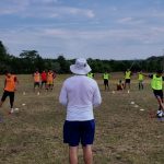 The soccer players racing through cones with a coach in the middle watching them with his back to the camera.