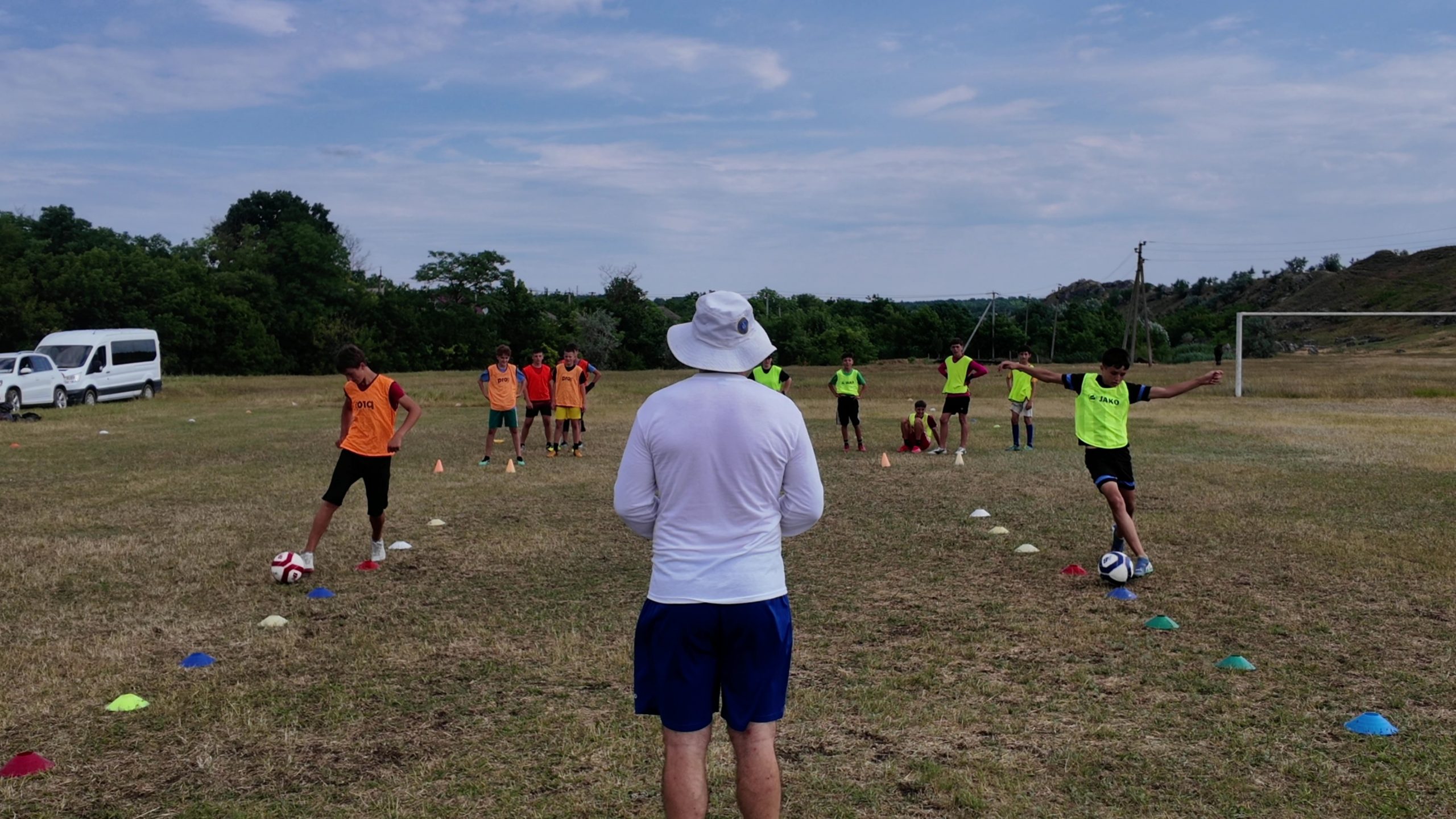 The soccer players racing through cones with a coach in the middle watching them with his back to the camera.